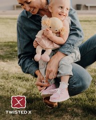 A woman and a young child crouching outdoors on grass, with the woman tying the child's pink shoes. The child is wearing a light pink dress and holding a doll.